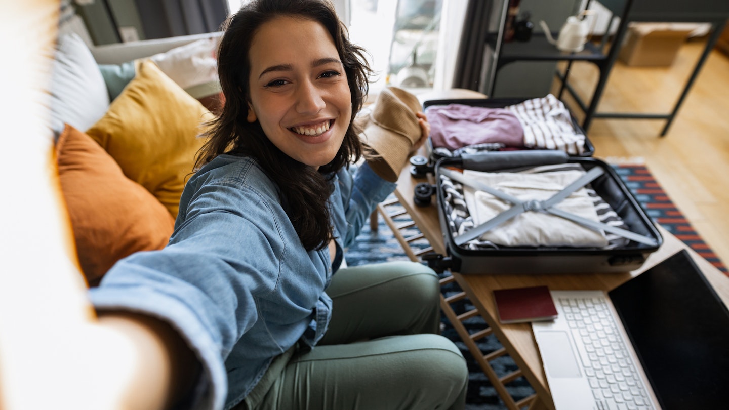 Young woman packing her suitcase and talking on a video call with her friends