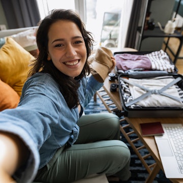 Young woman packing her suitcase and talking on a video call with her friends