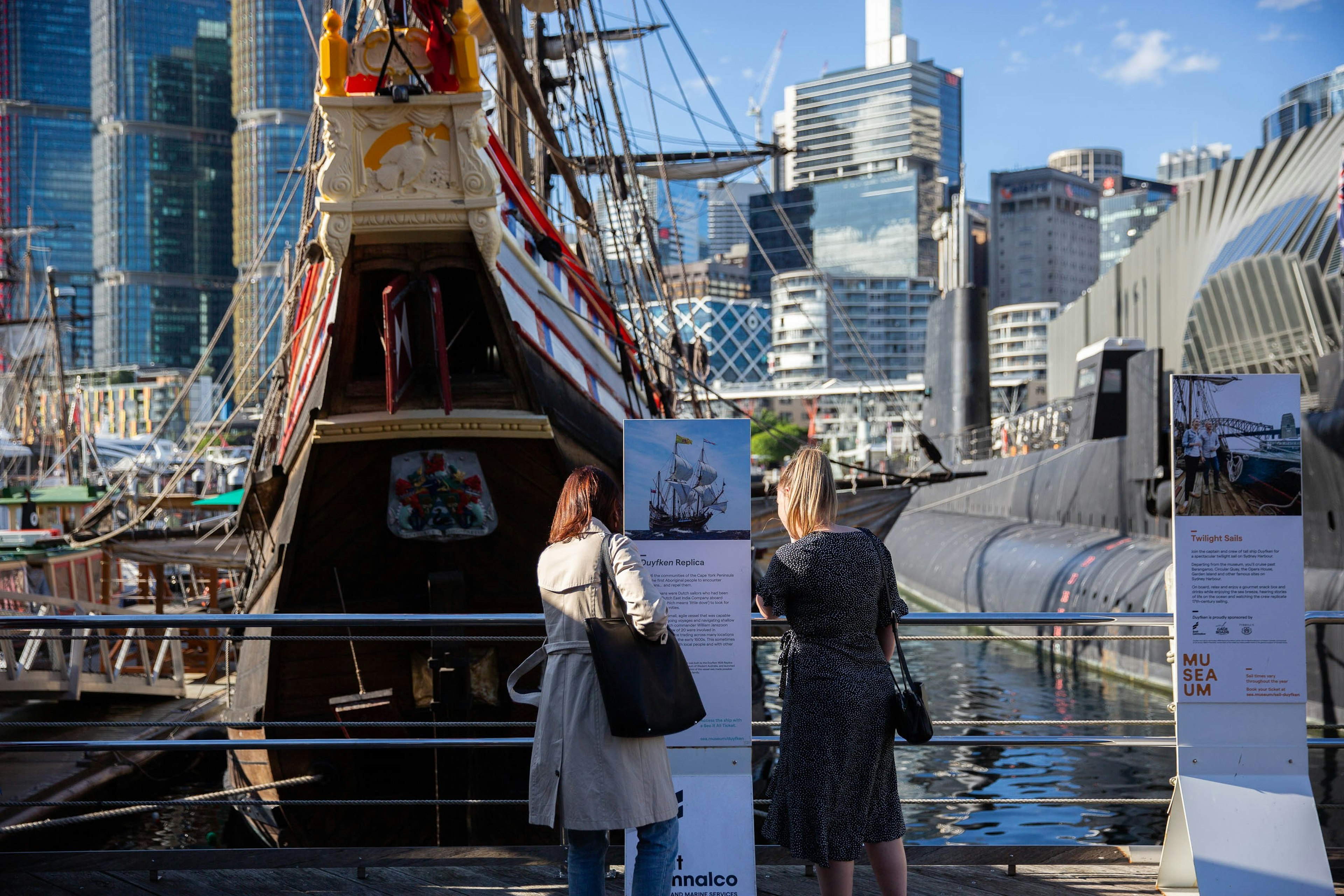 Two women stand on a dock in front of a an old wooden ship outside the Australian National Maritime Museum