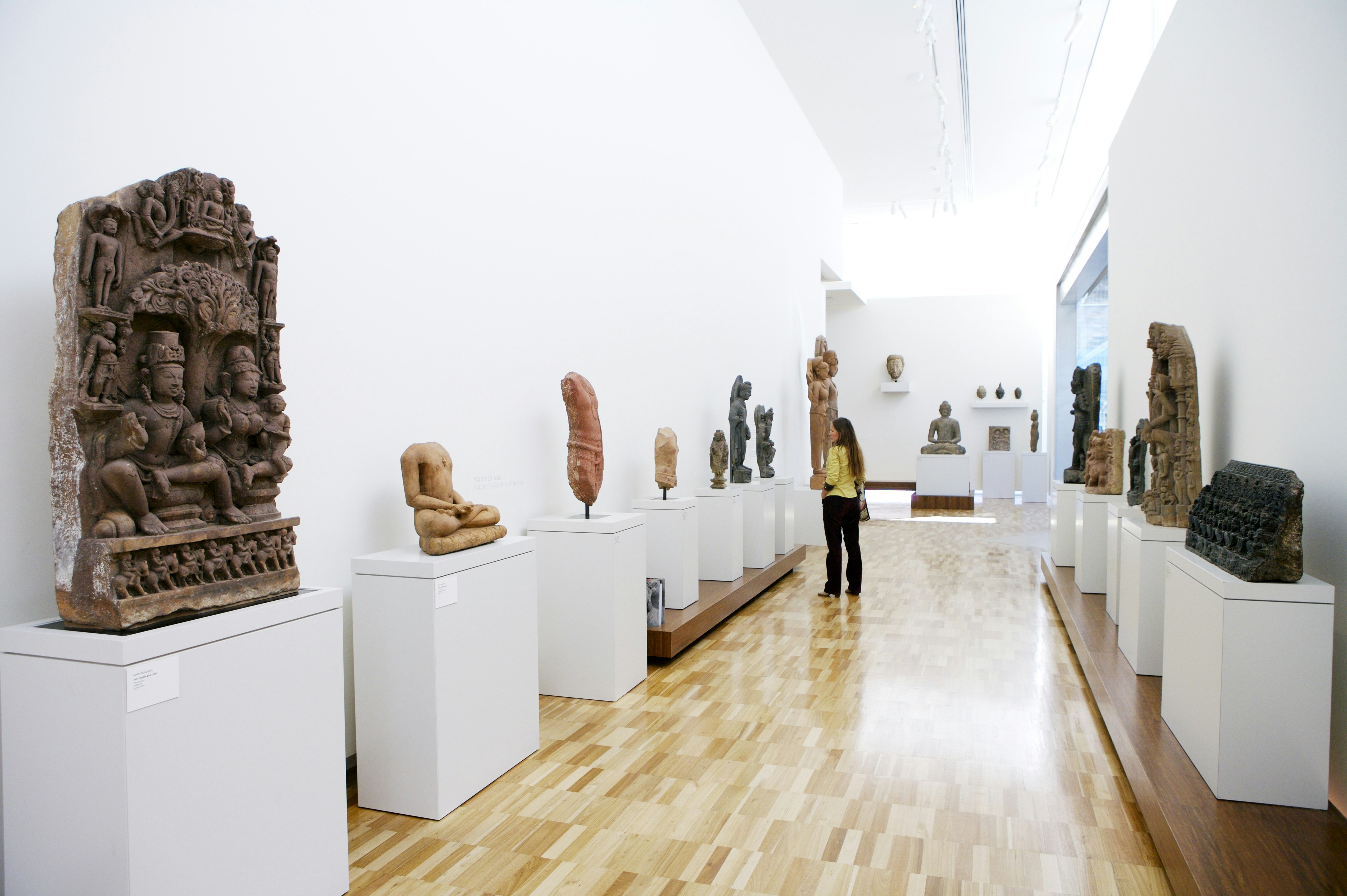 View of a long hallway with sculptures on either side. A woman is looking at a sculpture on one side of the hallway.