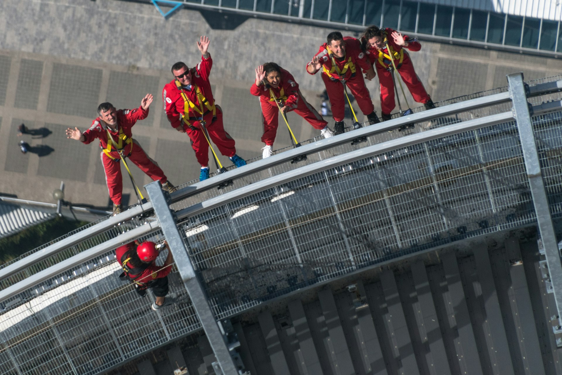 People in matching red jumpsuits dangle over the edge of a very tall building secured by yellow harnesses