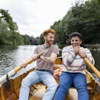 A male couple spending the day in Durham, England together. They are sitting in a rowboat and using one oar each to row the boat along the river. One man is looking at his fiancé and smiling while they row.