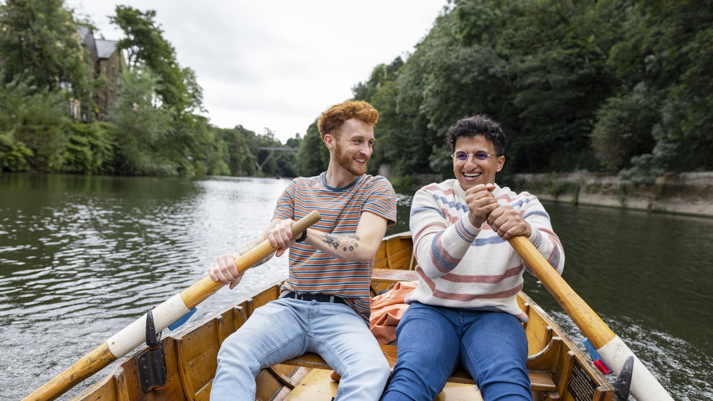 A male couple spending the day in Durham, England together. They are sitting in a rowboat and using one oar each to row the boat along the river. One man is looking at his fiancé and smiling while they row.