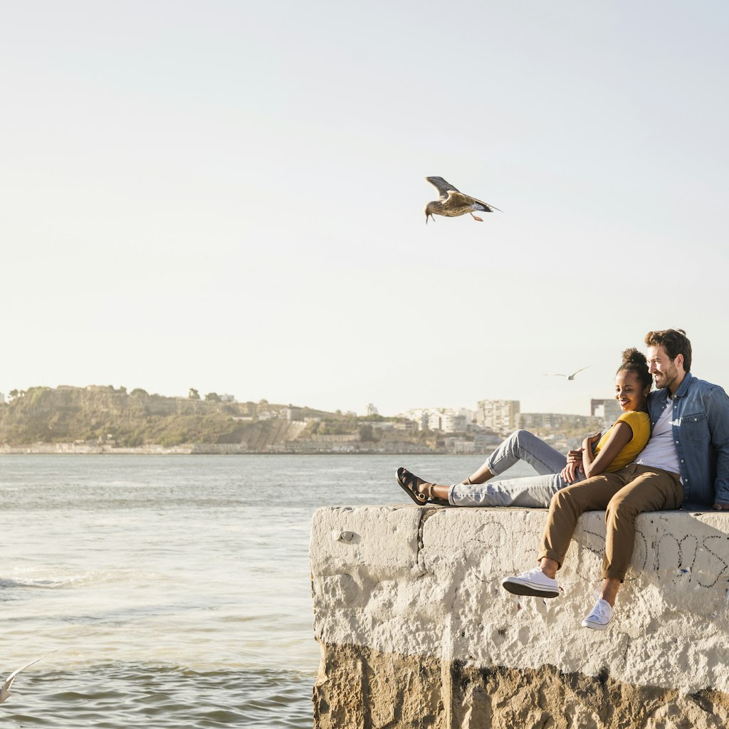 Young couple sitting on pier at the waterfront enjoying the view, Lisbon, Portugal