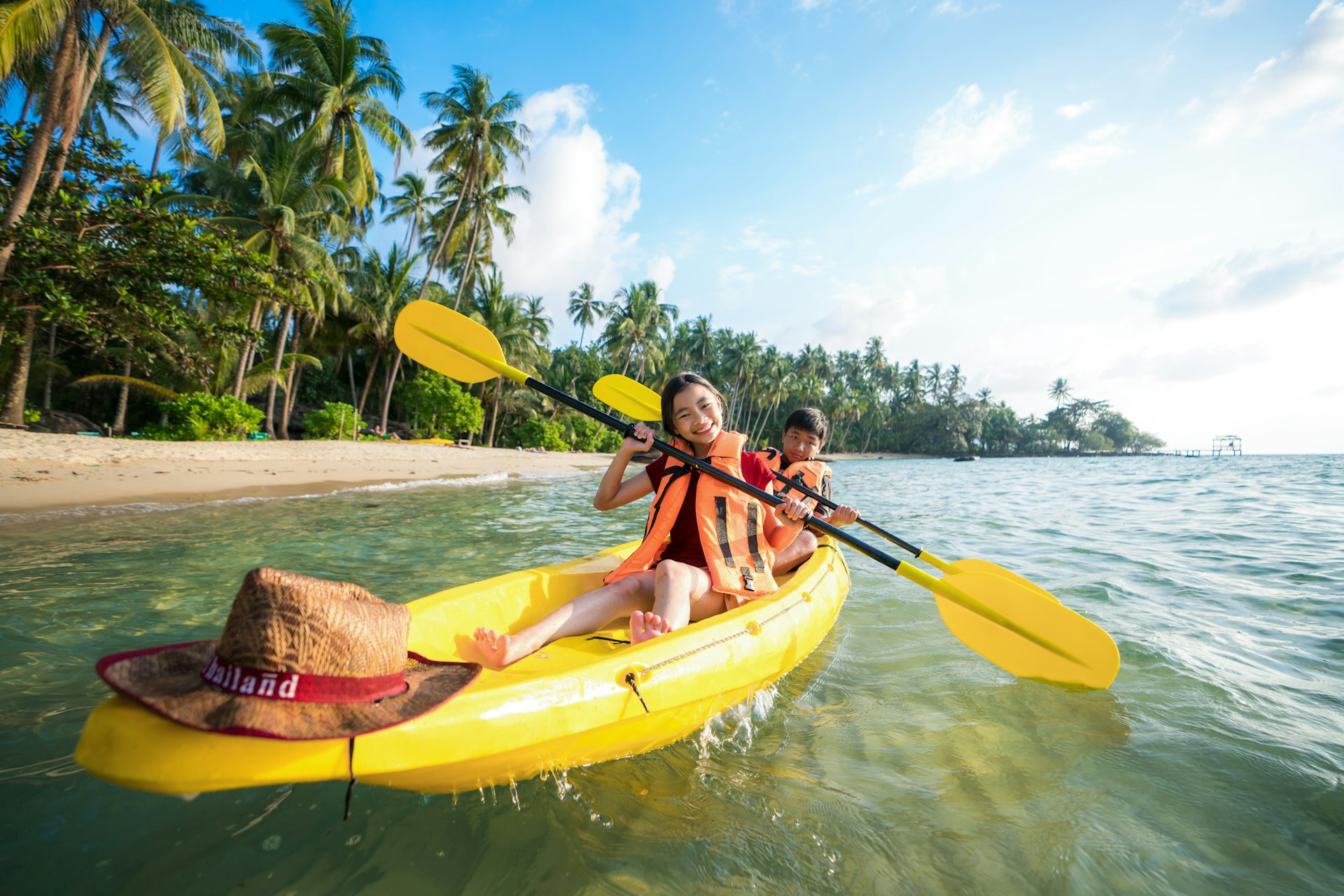 Two pre-teen children in a yellow kayak along a tropical palm-tree lined coastline