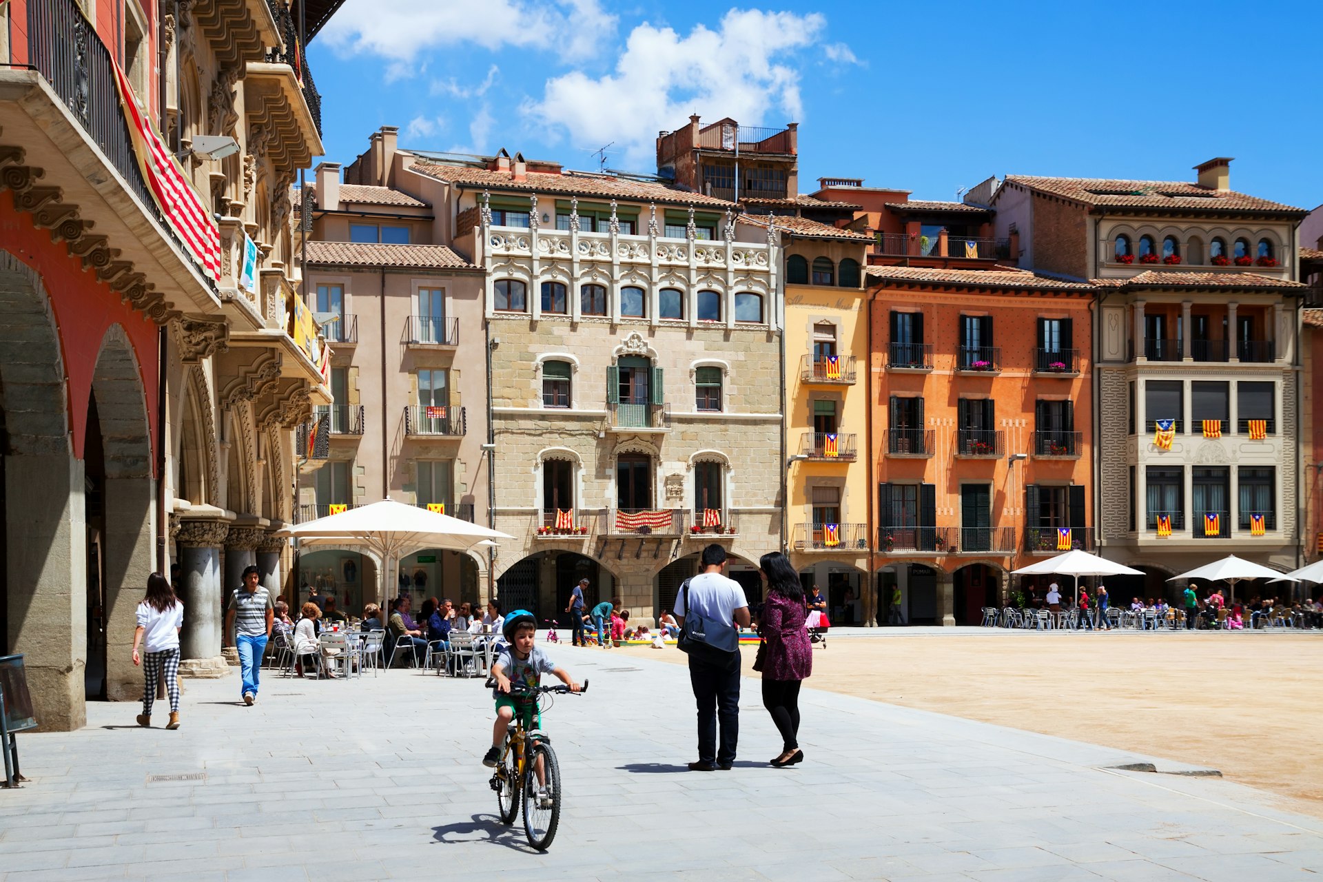 A child rides a bike and people walk around Plaça Mayor in Vic, Spain