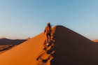 Young woman with long hair in hat and jumpsuit walking to the top of the big dune enjoying the beautiful sunrise at the remote desert in Namib-Naukluft National park, Namibia, Southern Africa