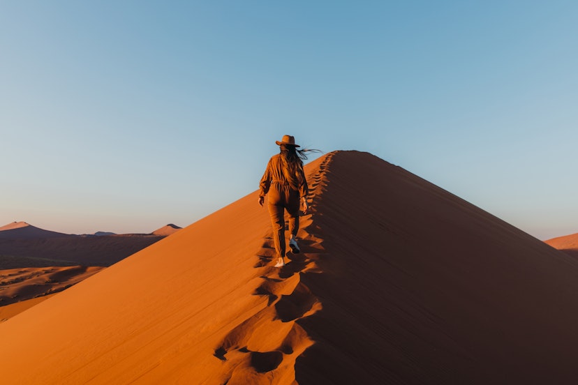 Young woman with long hair in hat and jumpsuit walking to the top of the big dune enjoying the beautiful sunrise at the remote desert in Namib-Naukluft National park, Namibia, Southern Africa