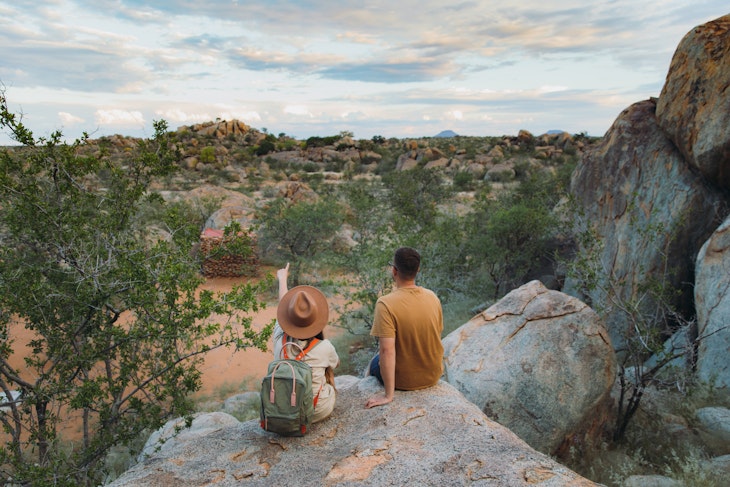 Young couple of woman in hat with backpack and man sitting on the top of the rock looking at the beautiful landscape and watching wild animals in Damaraland, Namibia, Southern Africa