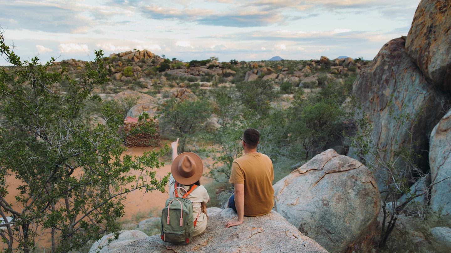 Young couple of woman in hat with backpack and man sitting on the top of the rock looking at the beautiful landscape and watching wild animals in Damaraland, Namibia, Southern Africa
