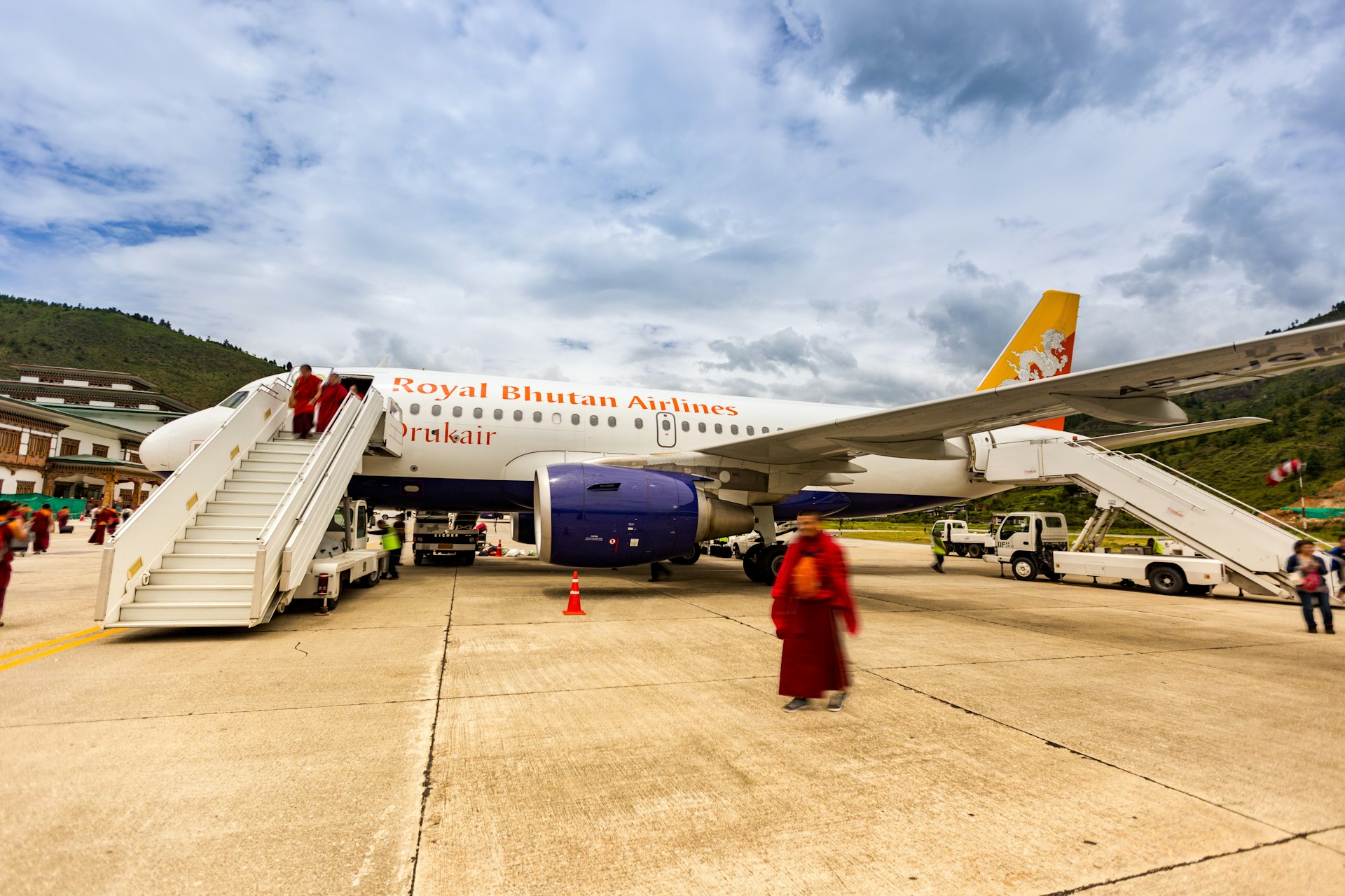 This daylight photo shows passengers arriving at Paro International Airport, Bhutan on a Royal Bhutan Airlines (Drukair) flight. The aircraft is an Airbus A319 with registration A5-JSW. A blur has been applied to all people in the photo. 