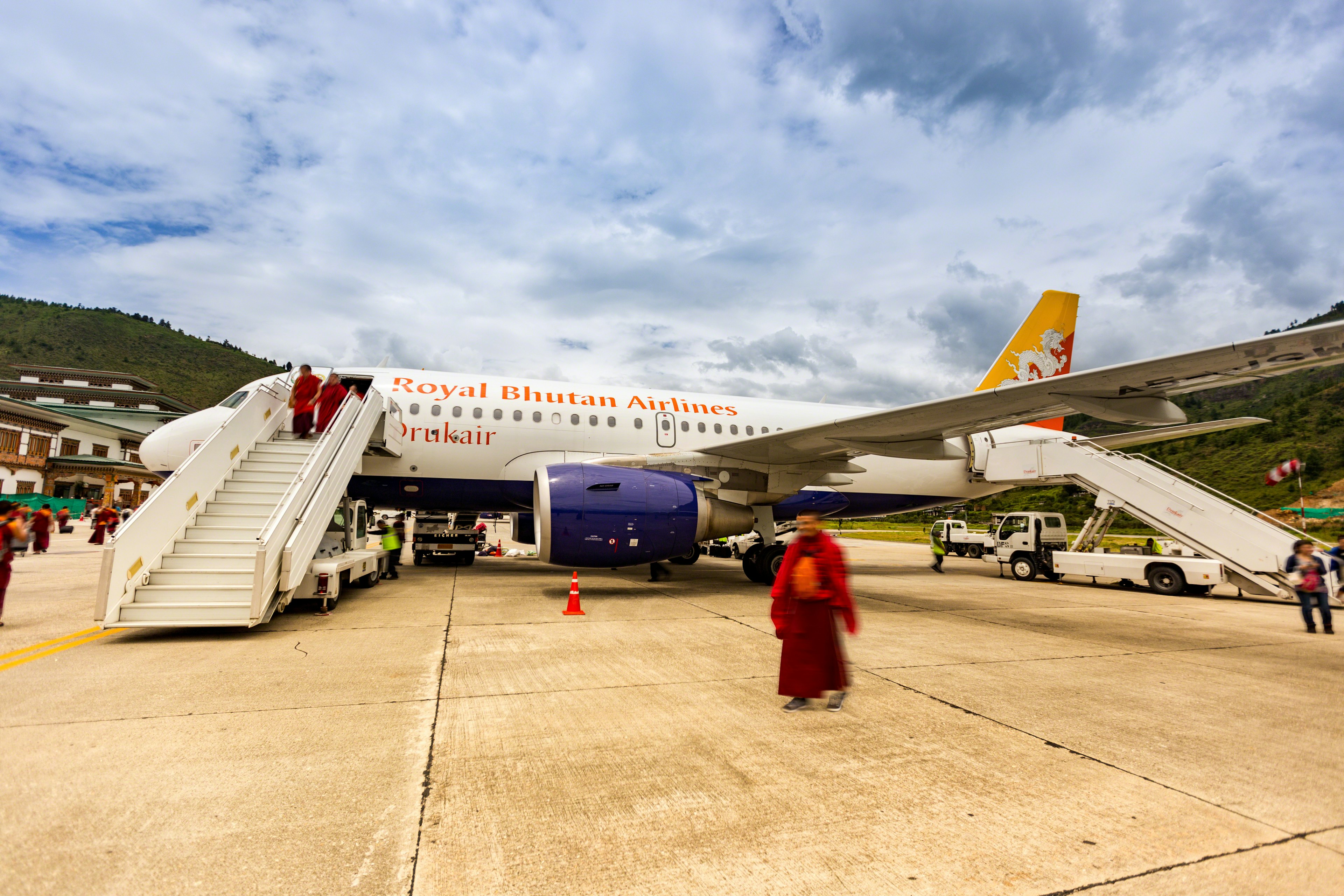 This daylight photo shows passengers arriving at Paro International Airport, Bhutan on a Royal Bhutan Airlines (Drukair) flight. The aircraft is an Airbus A319 with registration A5-JSW. A blur has been applied to all people in the photo.