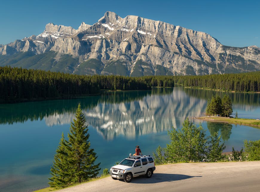 A car stopped by a lake in a mountainous landscape. A person sits on the roof taking in the view. 