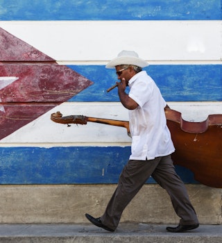 A musician walking in the street in front of a Cuban flag