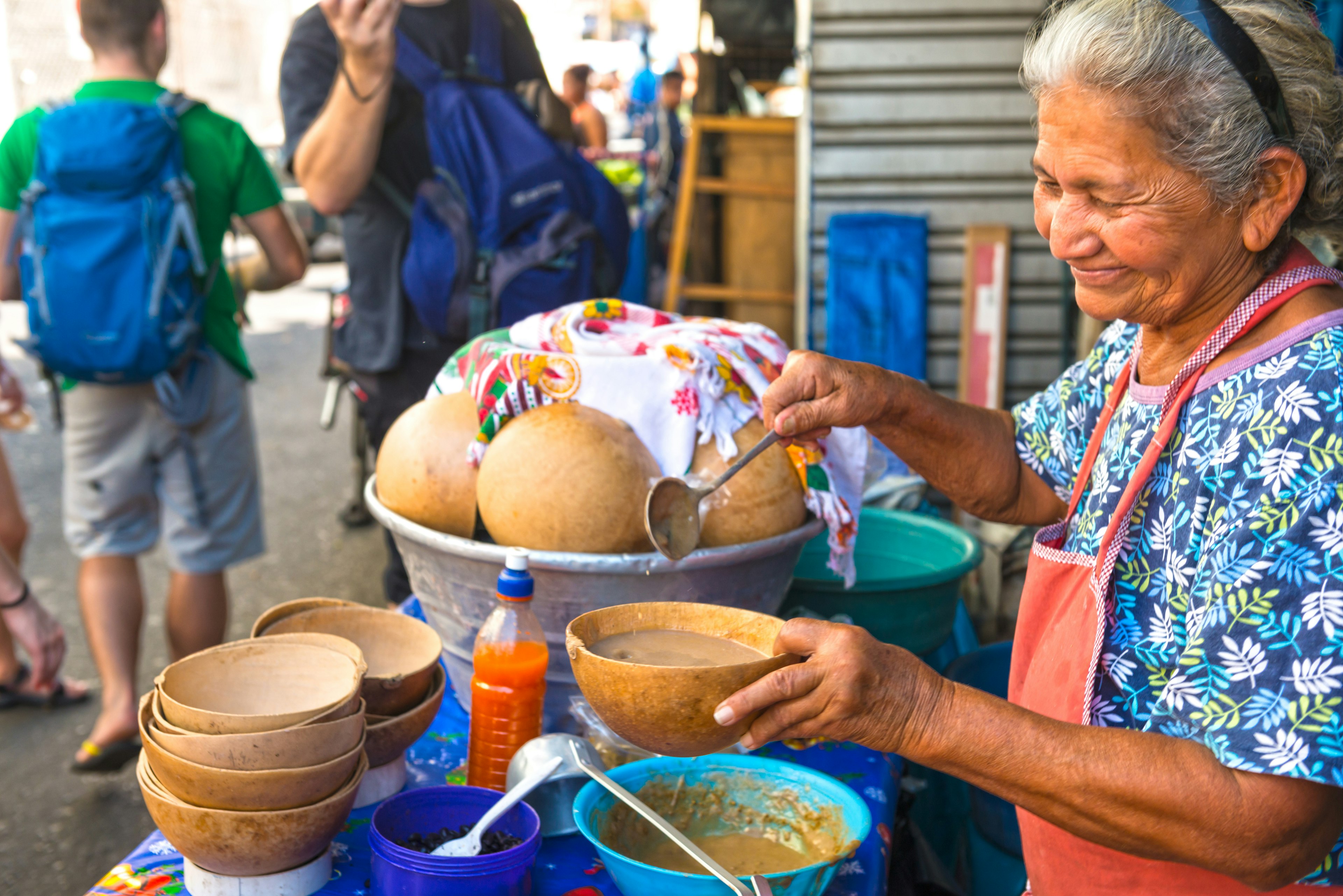 In front view old smilling woman pouring soup into the pumpkin bowl and selling. She has a food stall on the street. People in background.