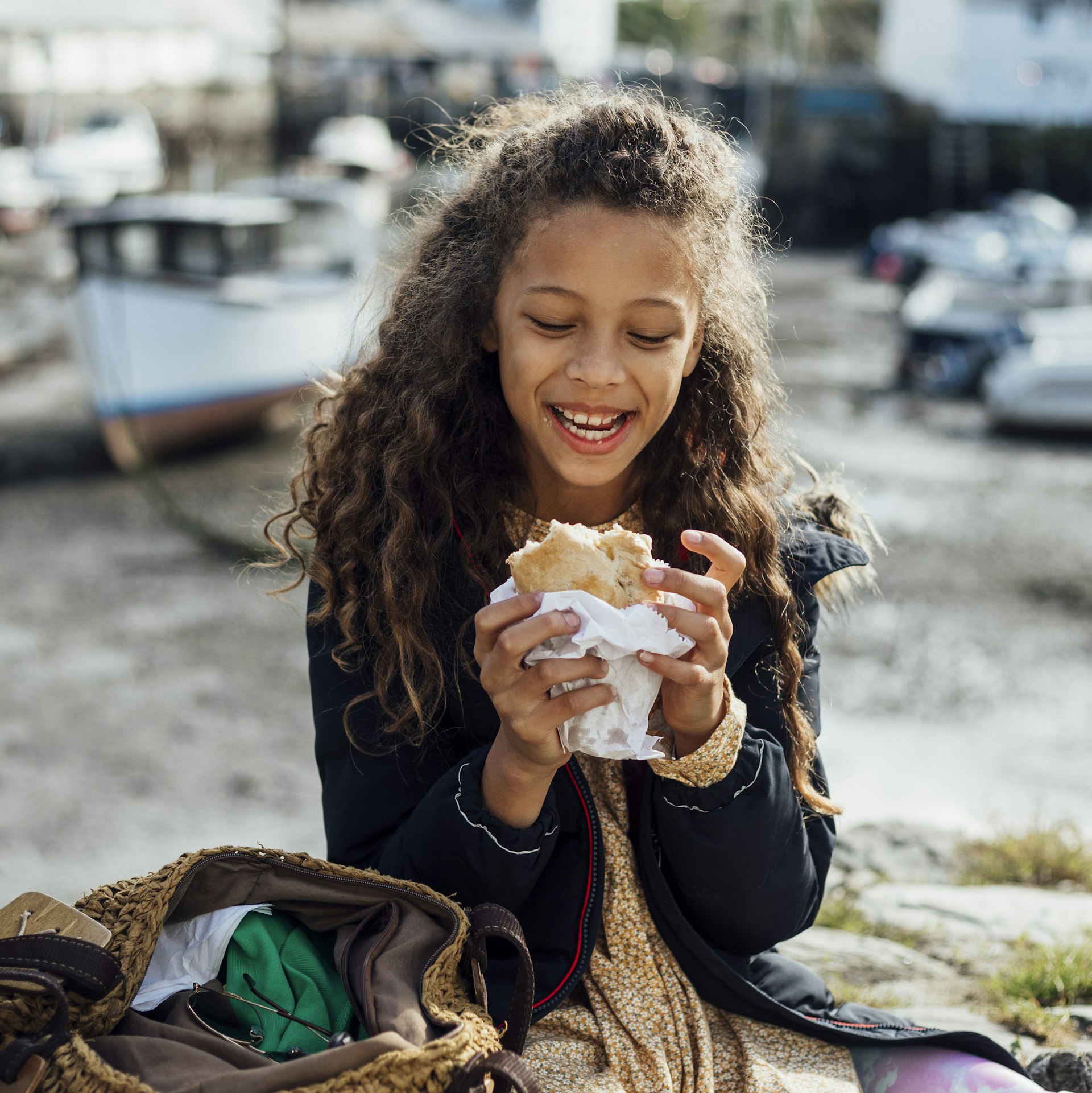 A young girl sitting on a stone wall in the fishing village of Polperro, Cornwall. She is eating a Cornish pasty while smiling.