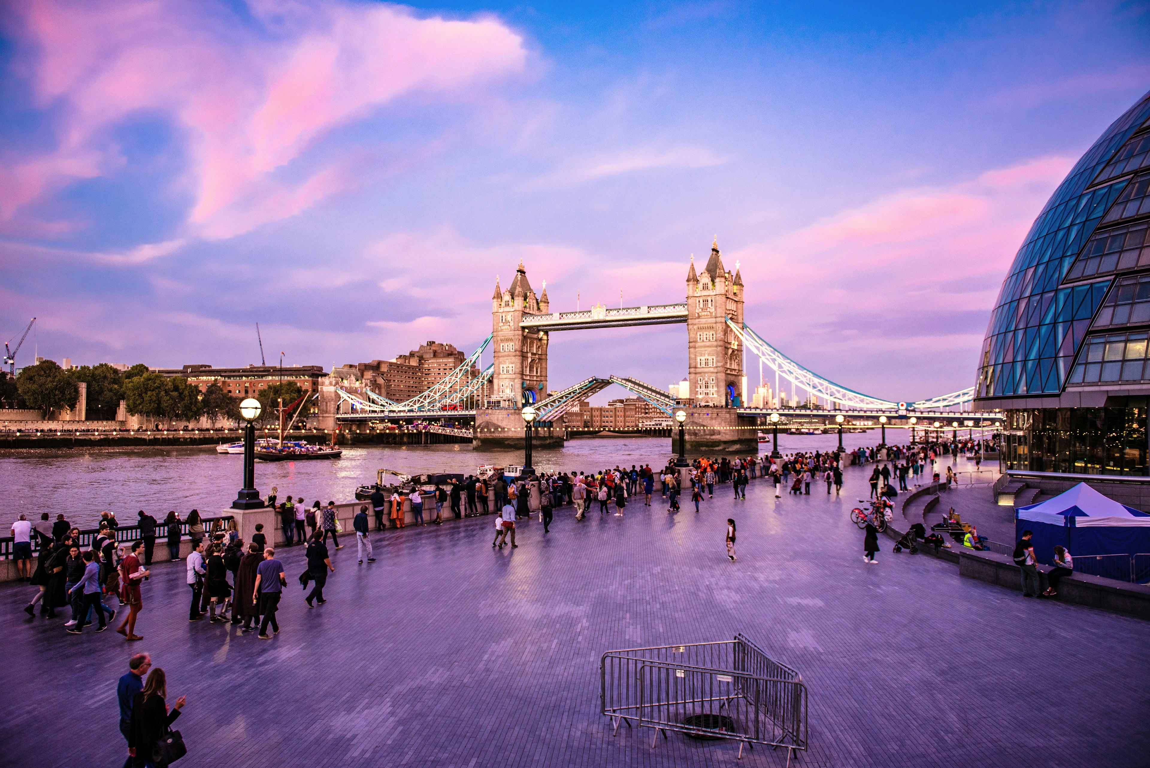 People mill around on the riverfront near a large Gothic bridge lit up in the evening