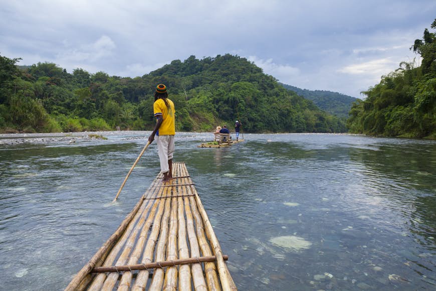 A pilot with a large pole guides a bamboo raft down a wide river