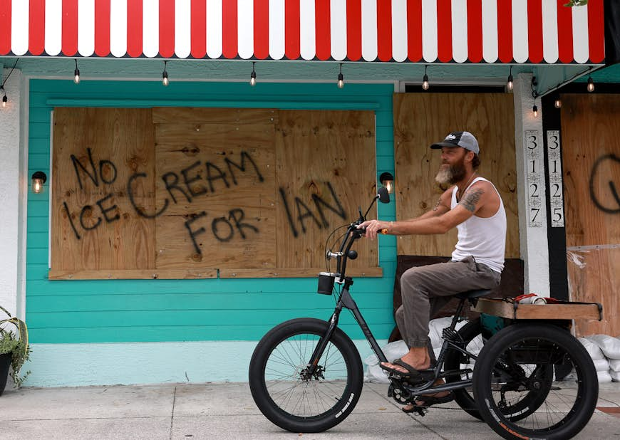 When a cyclist reads a sign that says 'No Ice Cream for Ian', he rides a boarded-up building in anticipation of Hurricane Ian. 