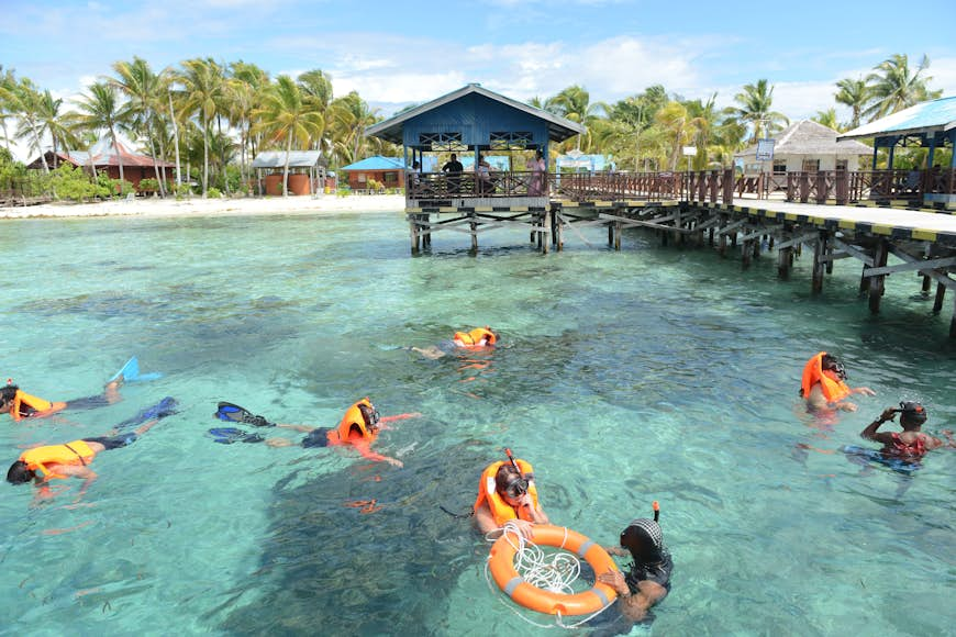 Snorkelers in life jackets look into the turquoise ocean just off shore from a settlement with small wooden houses 