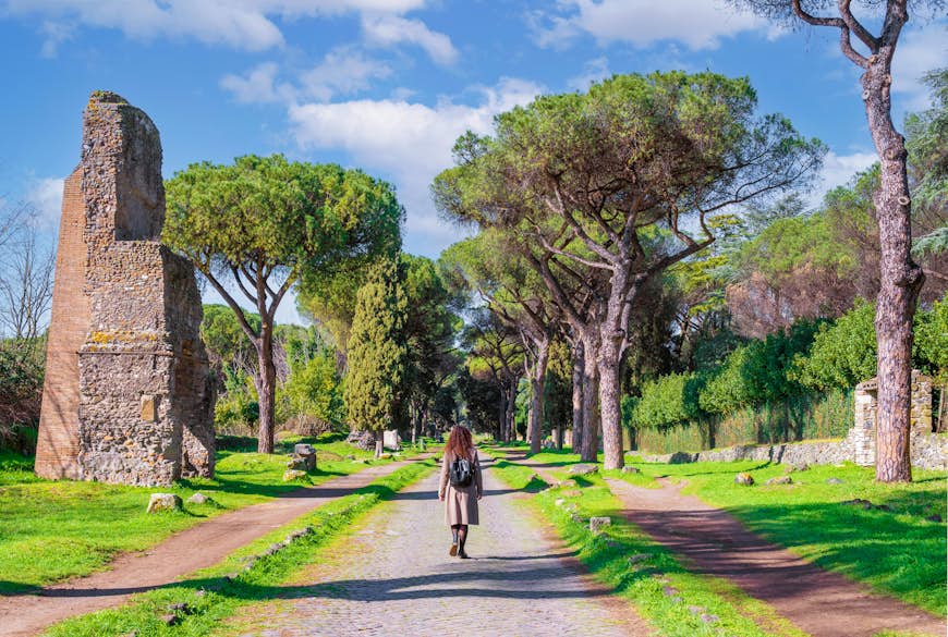 A solo figure walks down a tree-lined pathway on the Via Appia Antica, Rome