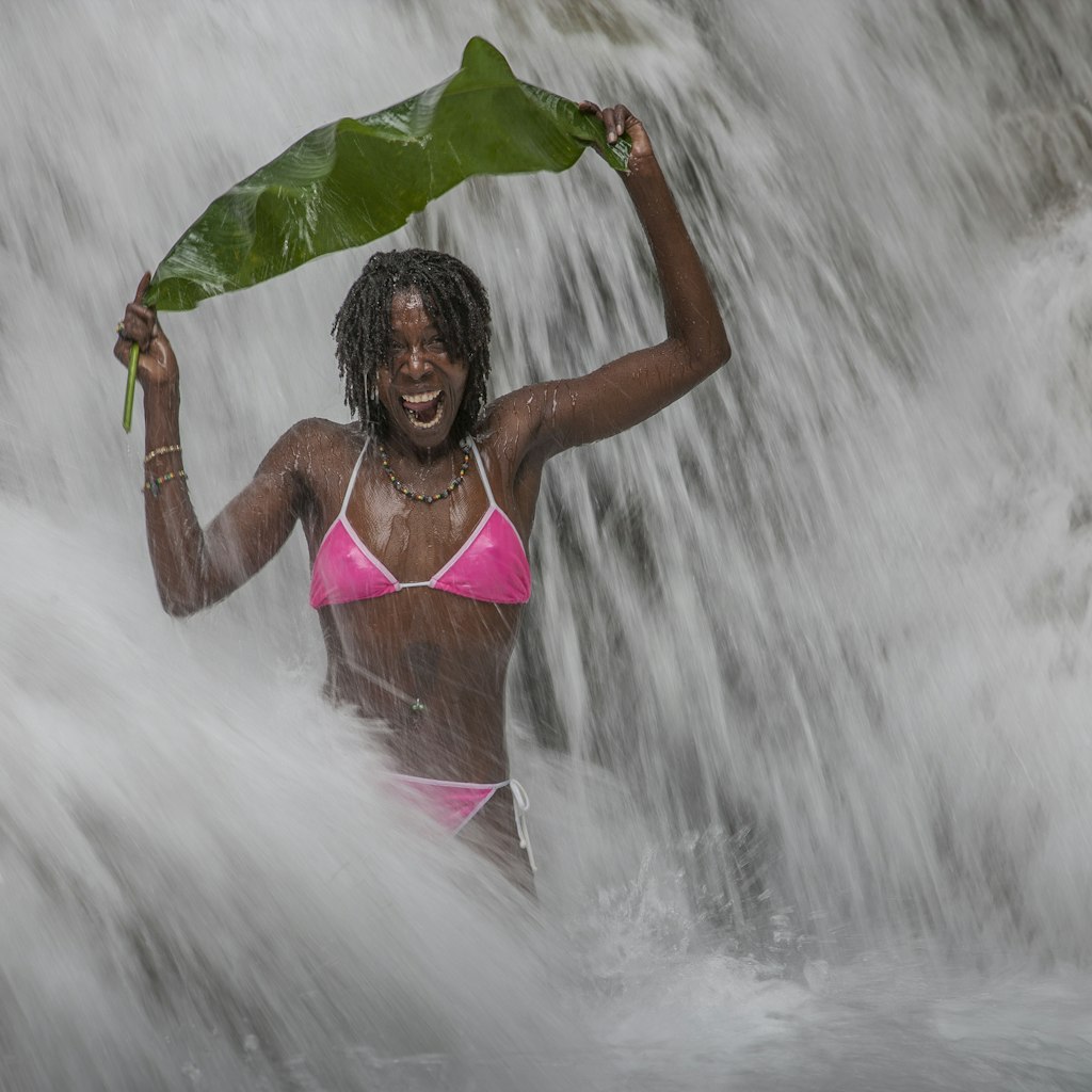 Jamaica. Echo Rios. Young smiling jamaican woman with pink bikini in the middle of the world famous Dunns River Falls, covering herself with a banana leaf.