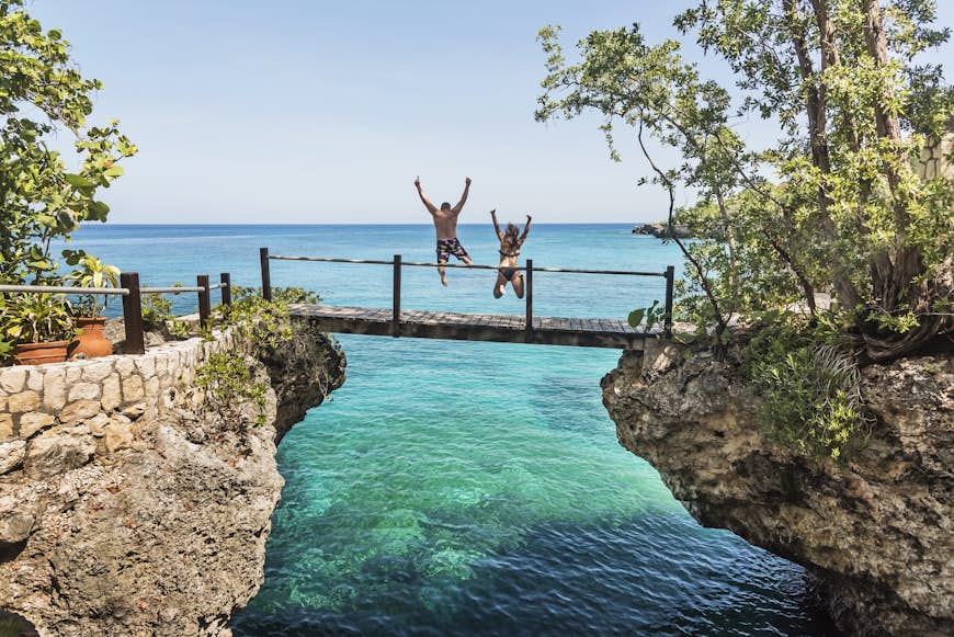 Two people in swimwear jump off a footbridge into the turquoise ocean