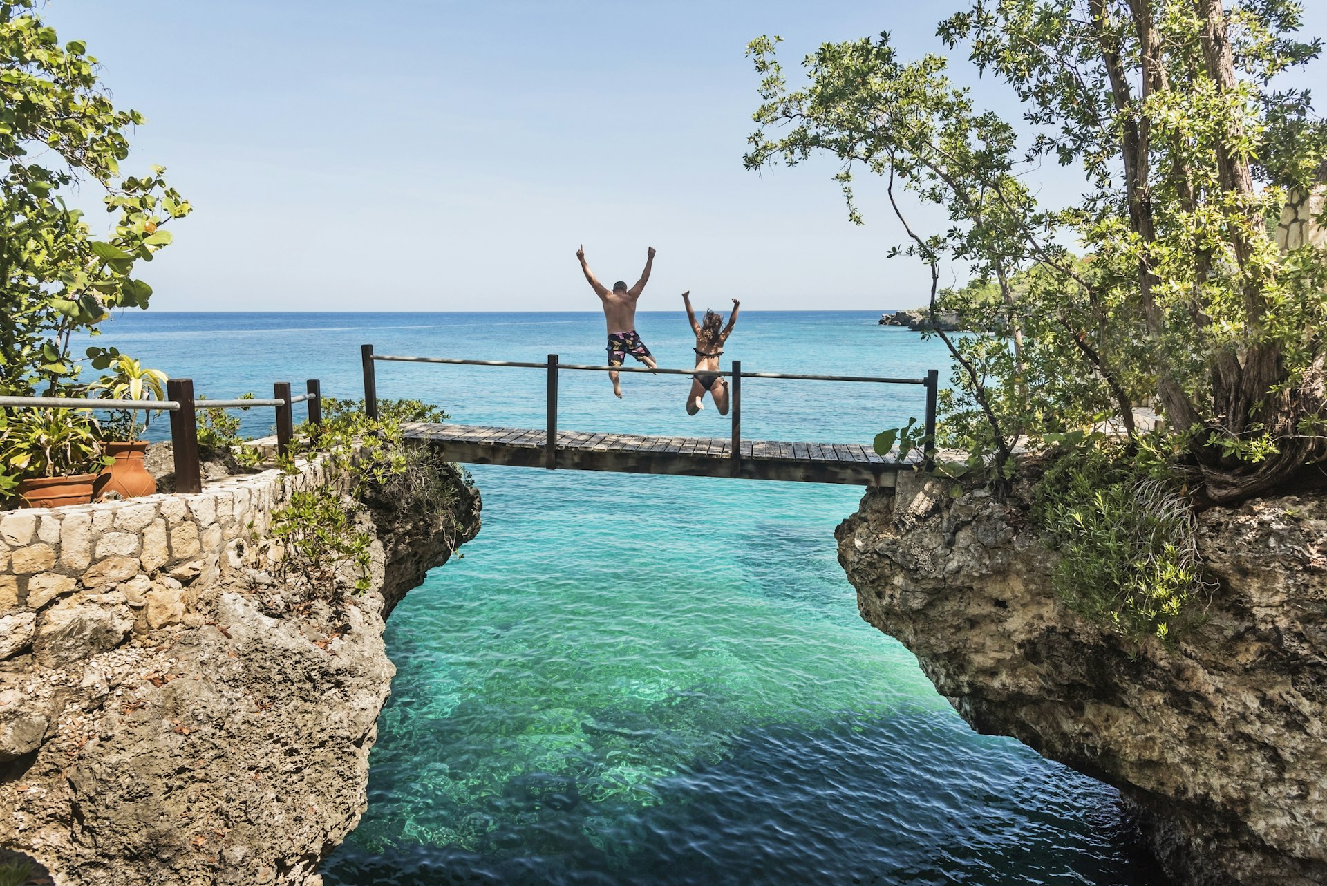 Two people in swimwear jump off a footbridge into the turquoise ocean