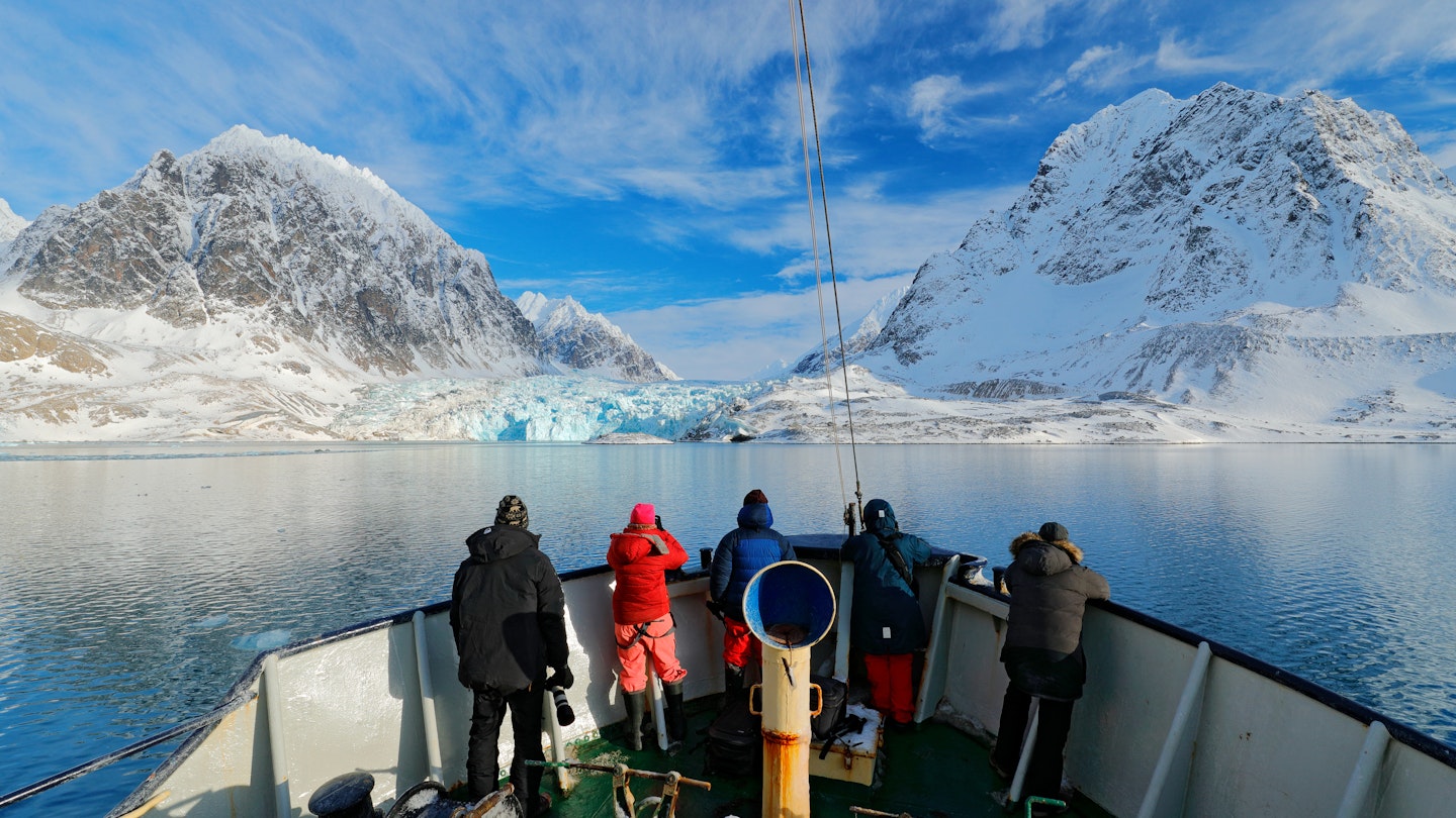 Holiday travel in Arctic, Svalbard, Norway. People on the boat. Winter mountain with snow, blue glacier ice with sea in the foreground. Blue sky with white clouds. Snowy hill in ocean. Travel in sea.