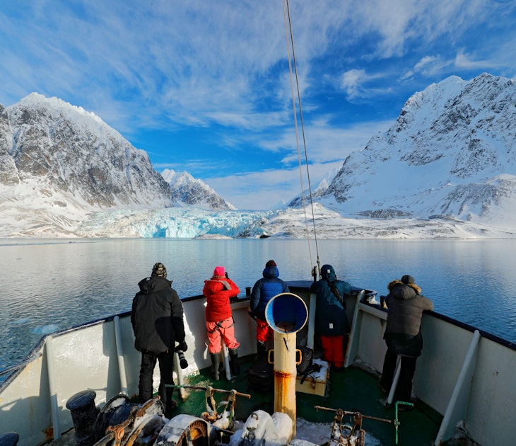 Holiday travel in Arctic, Svalbard, Norway. People on the boat. Winter mountain with snow, blue glacier ice with sea in the foreground. Blue sky with white clouds. Snowy hill in ocean. Travel in sea.