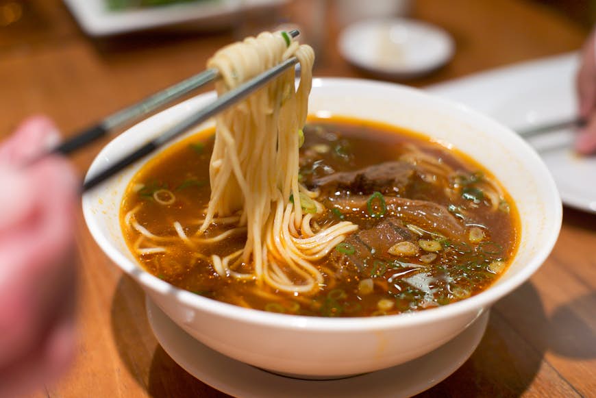 Someone lifts up some noodles from a bowl of brown Taiwanese beef noodles soup using chopsticks