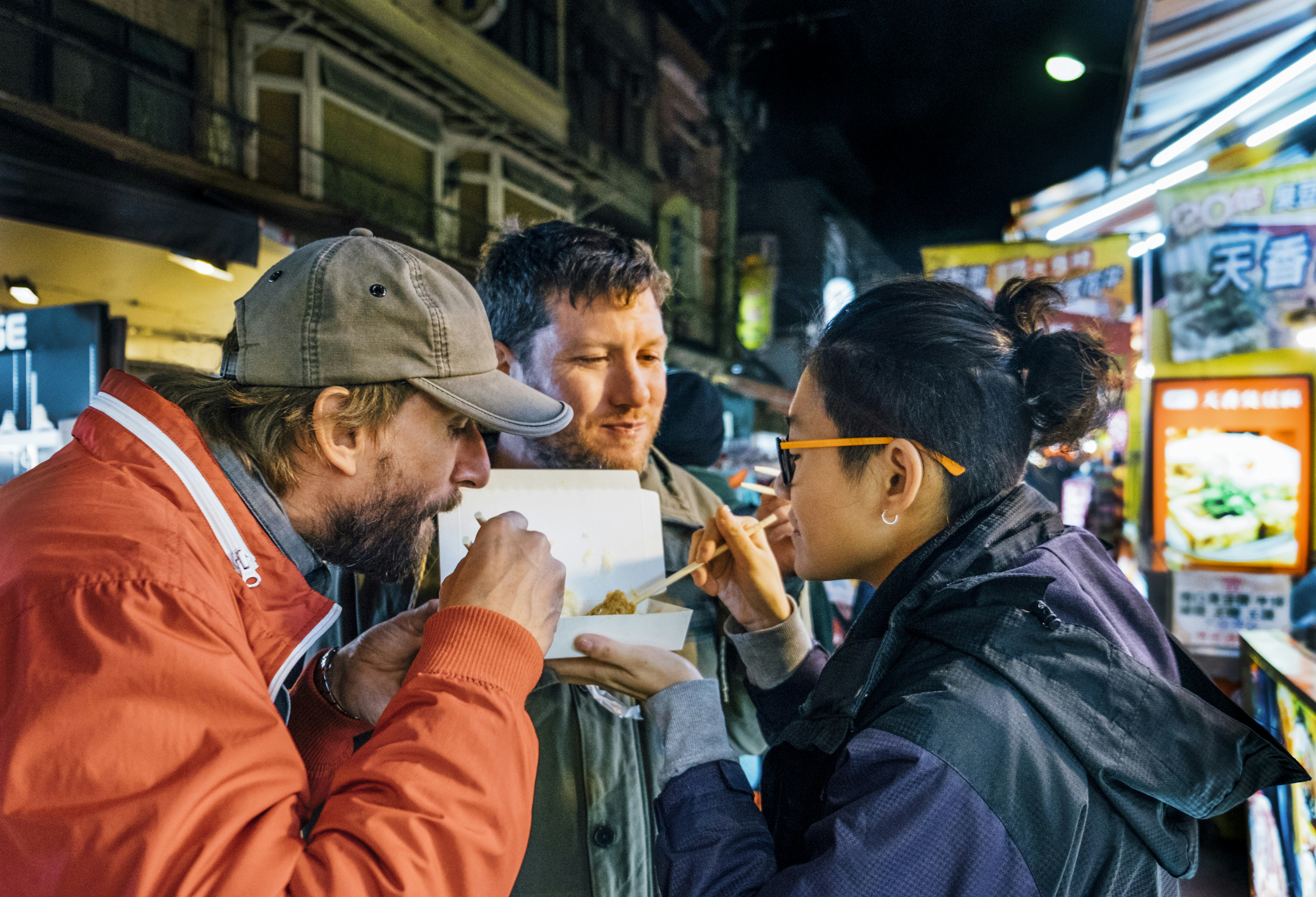 Two Caucasian males and an Asian female eat food at an outdoor Taiwanese night Market
