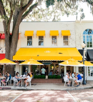 This is a horizontal, color photograph Park Avenue in downtown Winter Park, Florida. People dine at outside tables under umbrellas by the sidewalk.