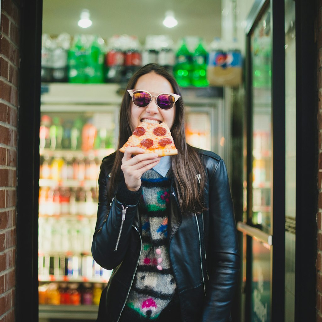 Happy young woman in sunglasses eating a slice of pizza from corner pizza joint in downtown New York City

