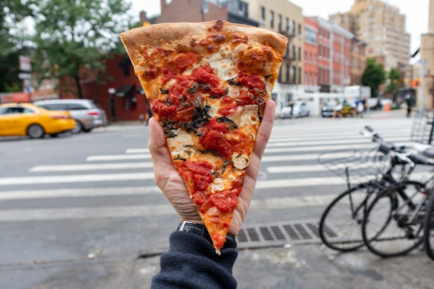 A New York style margherita pizza slice held by a hand along a New York City street 
