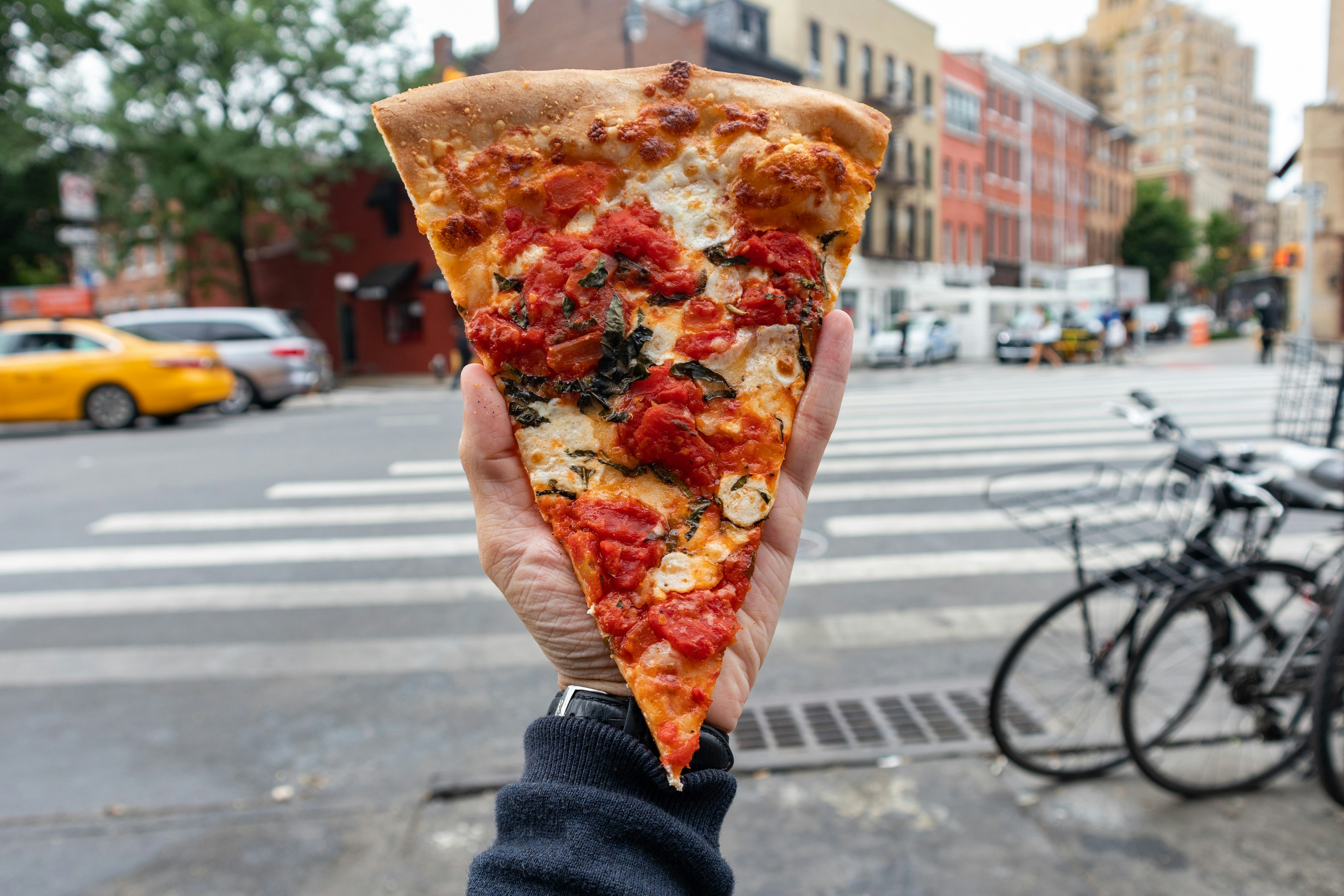 A New York style margherita pizza slice held by a hand along a New York City street