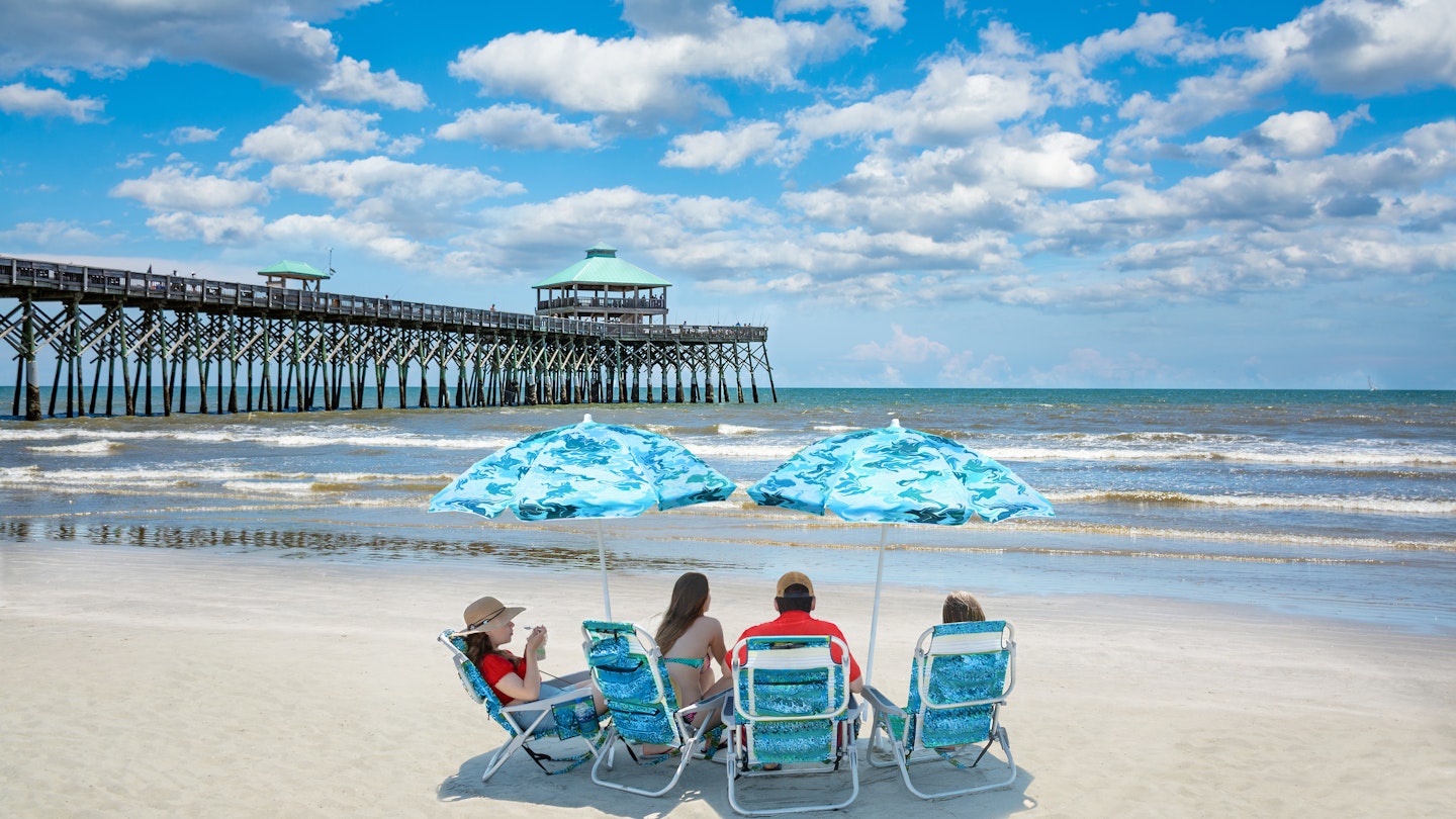 Family relaxing on  the beautiful beach, People enjoying summer vacation by the ocean. Family sitting under beach umbrella.  Cloudy sky and pier in the background. Folly Beach, South Carolina USA. ; Shutterstock ID 1428607988; your: Sloane Tucker; gl: 65050; netsuite: Online Editorial; full: Free Things South Carolina Article