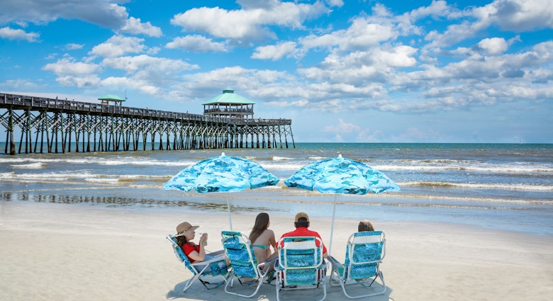 Family relaxing on  the beautiful beach, People enjoying summer vacation by the ocean. Family sitting under beach umbrella.  Cloudy sky and pier in the background. Folly Beach, South Carolina USA. ; Shutterstock ID 1428607988; your: Sloane Tucker; gl: 65050; netsuite: Online Editorial; full: Free Things South Carolina Article
