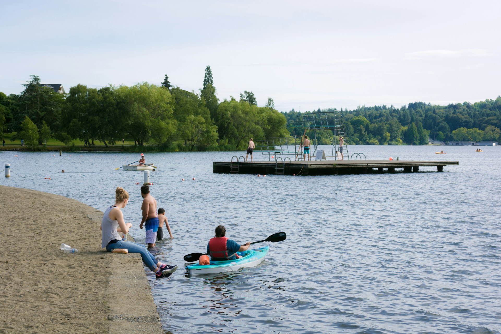 People are meander about the water's edge at Green Lake in North Seattle while others play on the dock in the distance or relax in water craft on the lake.