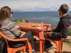 young white caucasian man and woman couple sitting in an outside restaurant on orange plastic chairs and table, eating lunch and enjoying the view vista viewpoint over lake titicaca on isla del sol, island of the sun, bolivia