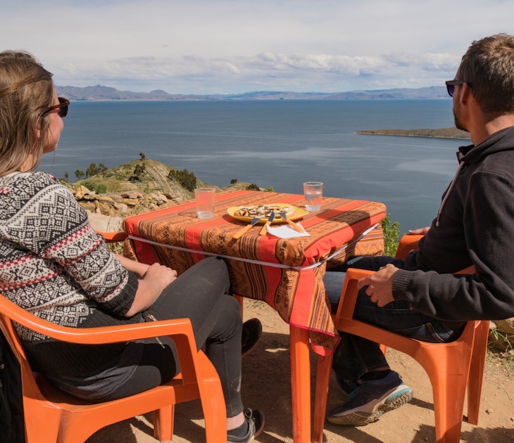 young white caucasian man and woman couple sitting in an outside restaurant on orange plastic chairs and table, eating lunch and enjoying the view vista viewpoint over lake titicaca on isla del sol, island of the sun, bolivia