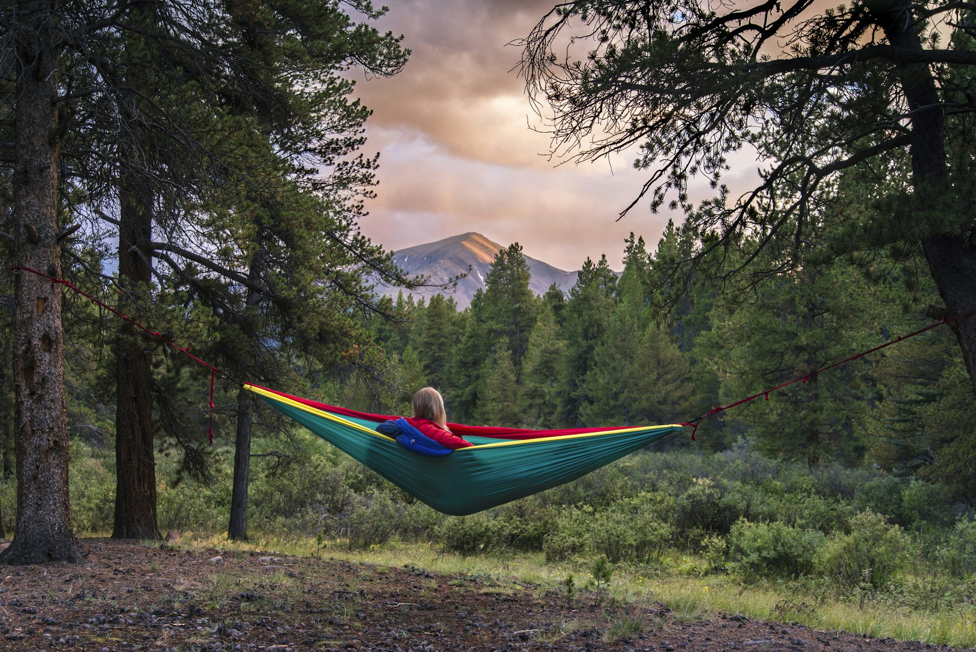 Woman sits in a colorful hammock in a forest in Colorado overlooking a mountain peak