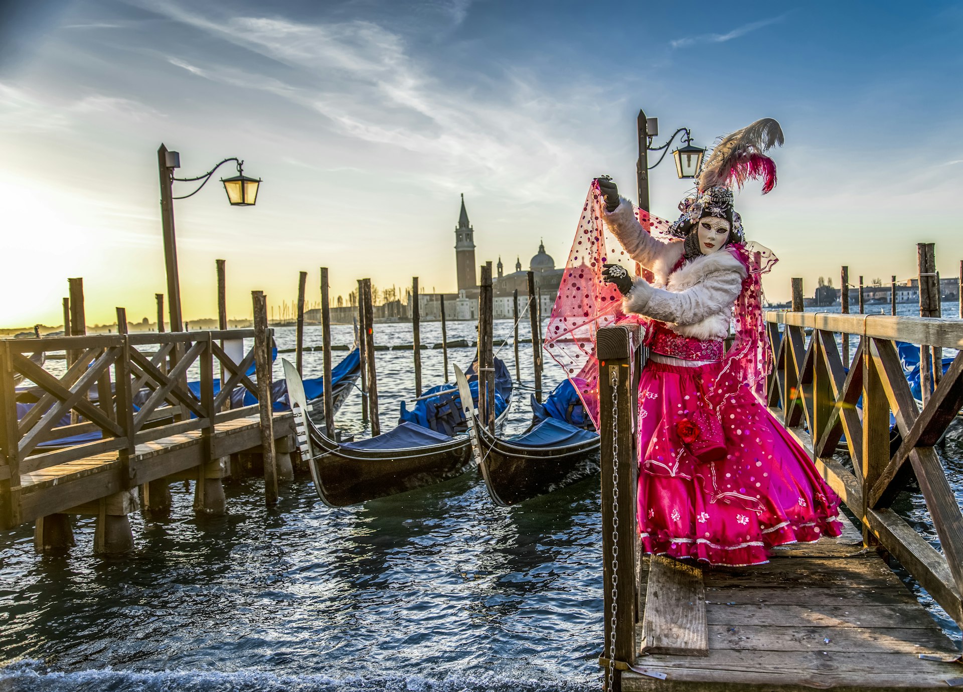 A masked person in a grand costume poses in front of gondolers docked at the side of a canal in Venice