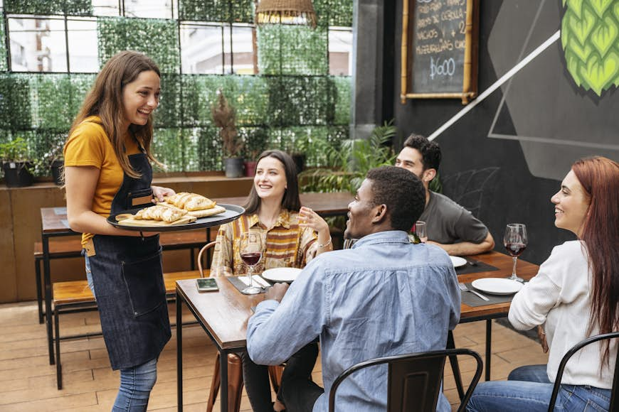 Smiling waitress holding cutting boards filled with fresh empanadas at table of four young African and Hispanic customers in Buenos Aires restaurant