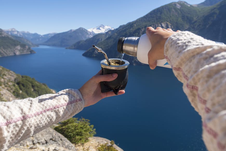 A tourist pours out a mate from a flask whilst looking out across a lake and mountains