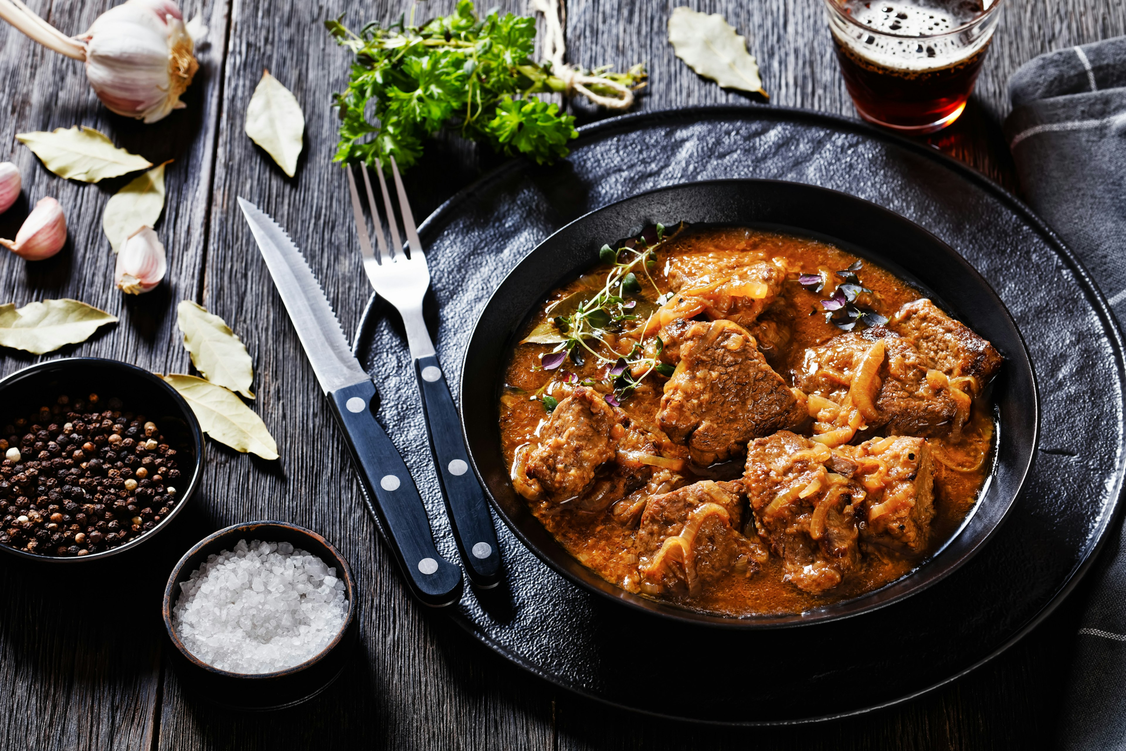 A black bowl is filled with a meaty stew, with pepper and utensils placed beside.