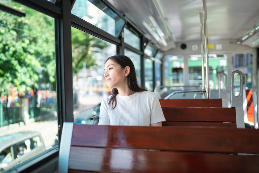 A smiling young tourist woman sits on double decker tram in Hong Kong on a sunny day