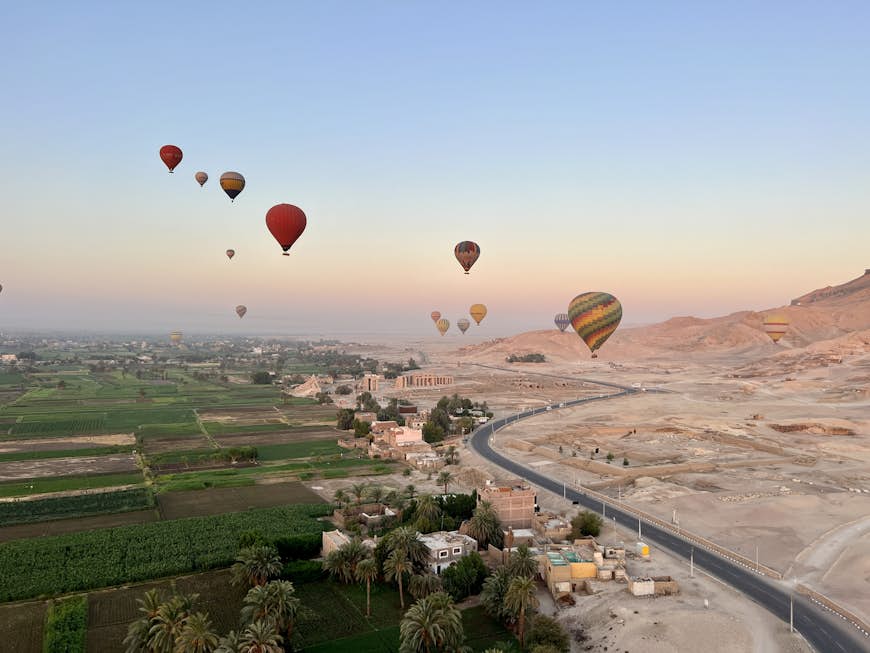 Colourful hot-air balloons fill the sky over a landscape of desert to the right and greenery to the left