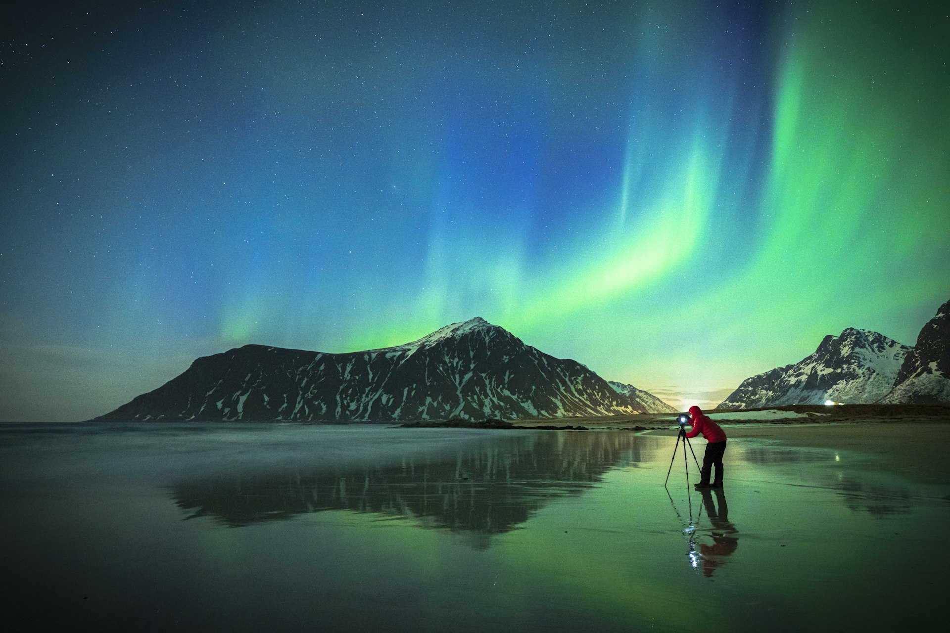 A photographer stands with their camera on the beach at night, photographing the northern lights. 