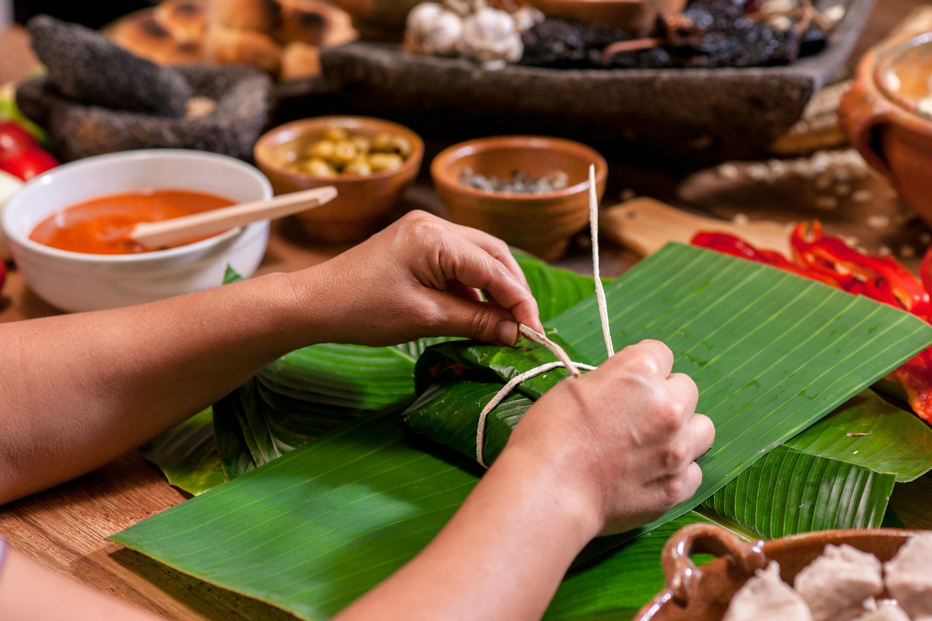 woman's hand tying and wrapping traditional Guatemalan tamales with banana leaves on a rustic wooden table with ingredients and spices at the workplace